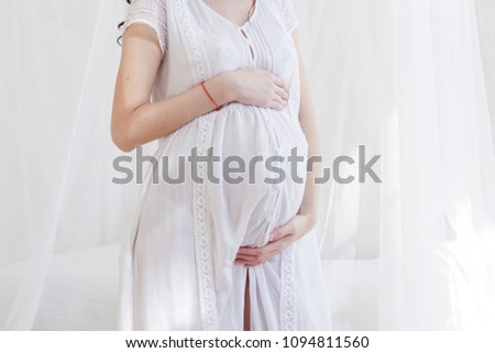 Pregnant woman in white dress holding her hands on her stomach. Closeup photo
