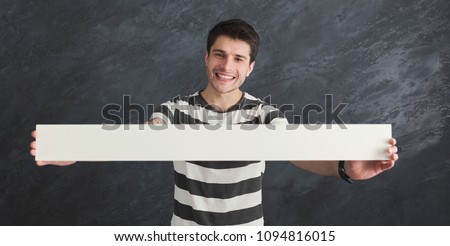Young tattooed man with blank white banner. Smiling man holding advertising sheet, gray studio background, copy space