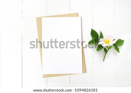 Feminine wedding stationery, floral desktop mock-up scene. Blank greeting card, craft envelope and blooming wild rose branch. Old white wooden table background. Flat lay, top view. 