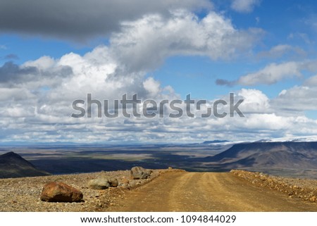 A gravel road leads over a small hill, behind it is a wide landscape with plain, mountain ranges and glaciers to see, above blue sky with white and gray clouds - Location: Iceland, highlands
