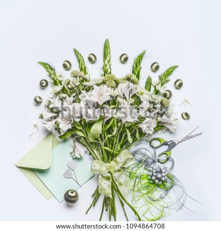 Beautiful festive bouquet  with green flowers on white desk background with envelope and paper card mock up, ribbon and scissors, top view. Holidays greeting concept