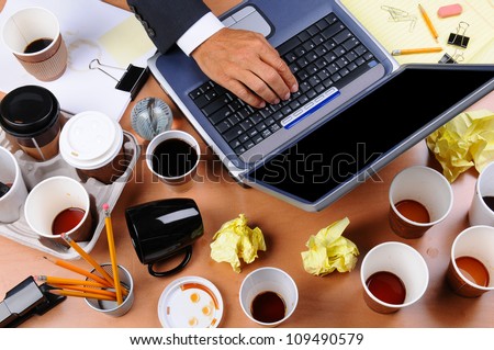 Closeup view of a very cluttered businessman's desk. Overhead view with man's hand on laptop keyboard and scattered coffee cups and office supplies. Horizontal format.