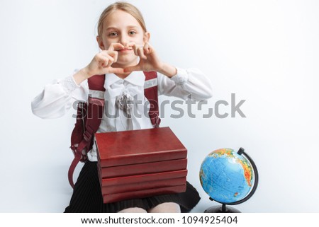 little cute schoolgirl girl sitting with books and globe, smiling and showing heart, white background, advertising,