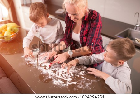 Mom and two sons are cooking in the kitchen. The flour is scattered on the table. A young mother in a red shirt is cooking in the kitchen.