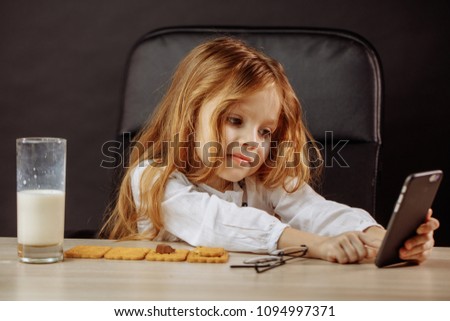 Children, modern technology and family relationships concept. Adorable little girl playing with smartphone in Daddy s office while having a snack with milk