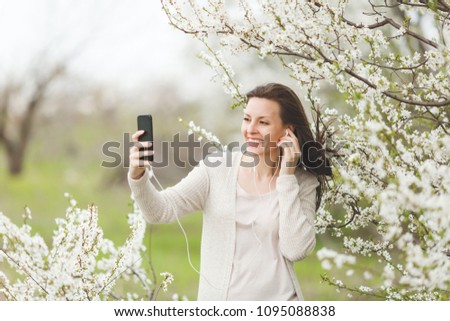 Young happy pretty woman in light casual clothes with earphones listening music doing selfie on mobile phone in city garden or park on blooming tree background. Spring flowers. Lifestyle concept