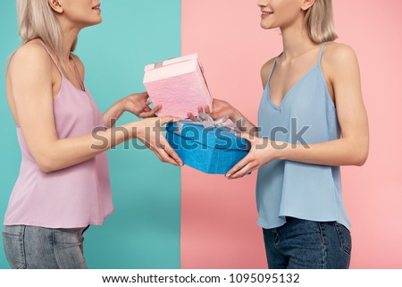 Two glad women standing one against another and exchanging present boxes. blue and pink background