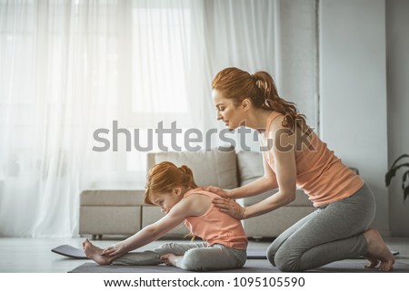 Full length portrait of woman helping her daughter to do stretching exercise on the floor. Lady is smiling and leaning back of her baby girl who looking concentrated