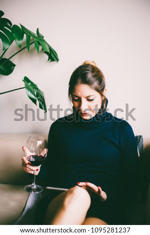 Beautiful young woman sitting on sofa, looking at tablet screen and drinks red wine (toned image, selective focus)