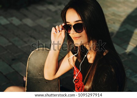 Close up portrait of cute long haired brunette sitting in a skatepark holding a skate and looking into camera smiling while listening music in headphones.