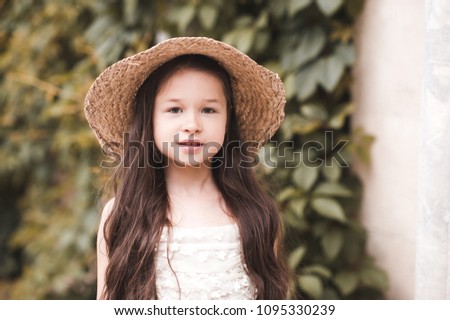 Smiling baby girl 4-5 year old wearing straw hat and white dress over nature background. Looking at camera. Summer vacation. 