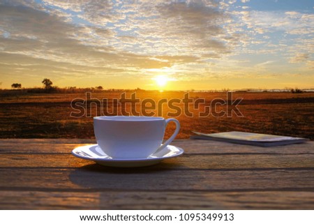 a selective focus picture of a cup of coffee on wooden table with organic green rice field in the morning sunrise