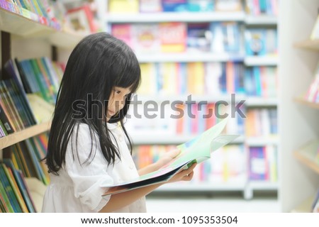 Asian children cute or kid girl smile reading and choose tale or story book on bookshelf in bookstore or library room at school or nursery for learning and studying with education