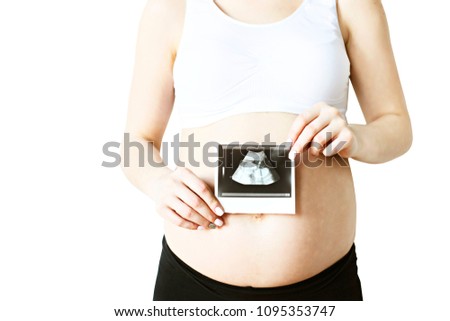 Close up of pregnant woman wearing supportive seamless maternity bra & black yoga pants holding fetus ultrasound scan of baby in her tummy, first photo of a child. White wall background.. Copy space.