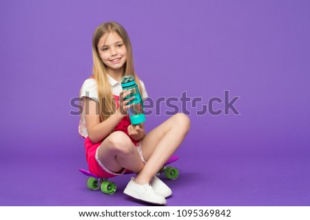Small girl sit on skate board with bottle on violet background. Child skater smile with water bottle. Skateboard kid in pink jumpsuit. Sport activity and energy. Drinking water, copy space.