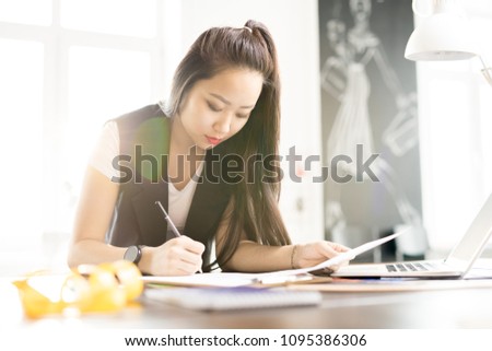 Portrait of creative young Asian woman drawing sketches for custom made clothes while standing at work table in sunlit fashion design atelier, lens flare