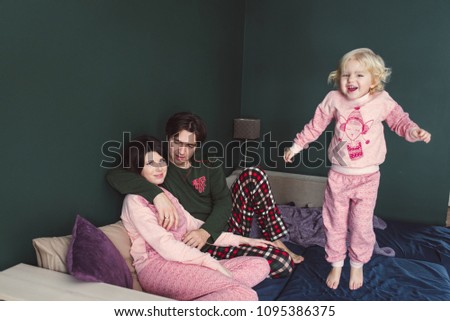 Mom and Dad are hugging sitting on the bed, and a little daughter is jumping on the bed. The little daughter jumps on the bed near her parents. Dad, mom and little daughter in beautiful pajamas.