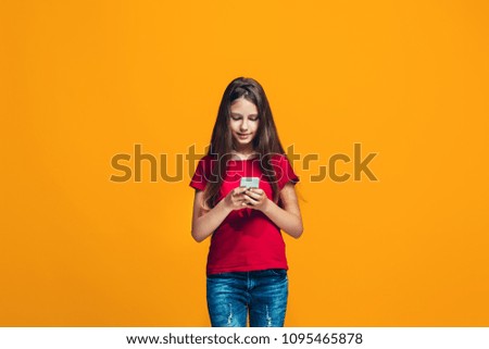 Happy teen girl standing, smiling with mobile phone over trendy orange studio background. Beautiful female half-length portrait. Human emotions, facial expression concept.