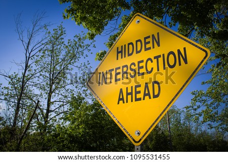 A yellow sign near trees indicating a hidden intersection ahead