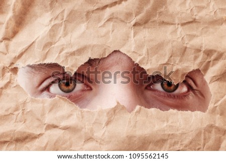 Eye looking through hole in paper. Spy eye watching through a hole. Eye looking through hole in old brown crumpled paper.