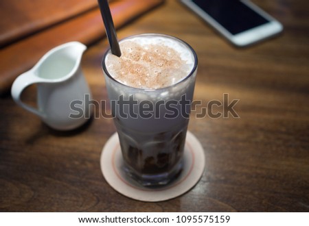 Ice Coffee with Milk Cream in Tall Glass with Dark Brown Straw inside and Mobile Phone, Leather Cover Book  on Wooden Table Vintage Style