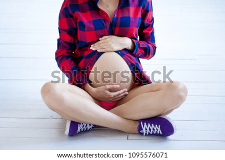 A pregnant woman sits on a white wooden floor. Purple sneakers and a red shirt in a cage. Royalty-Free Stock Photo #1095576071