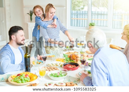 Portrait of happy family enjoying dinner sitting round festive table with delicious dishes, cute redhead girl hugging mom serving food during holiday celebration in modern sunlit apartment, copy space