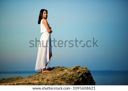 Young beatiful girl wearing white dress standing on rock near blue sea. Looking on amazing sideview, horizone. Feeling good, free, concentrated. Summer, warm, good weather.