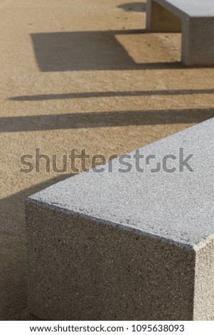 Close up outdoor view of two concrete benches in a public place, with their shadows drawn on the ground. Abstract design with parallelepiped shapes, oblique lines, grey, brown and white colors.  