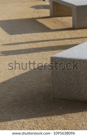 Close up outdoor view of two concrete benches in a public place, with their shadows drawn on the ground. Abstract design with parallelepiped shapes, oblique lines, grey, brown and white colors.  