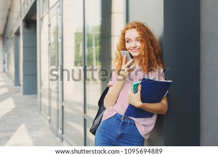 Attractive kind student girl with books on university background outdoors, looking away, talking on smartphone, having rest in campus. Education concept, copy space