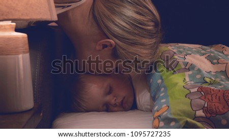 A little boy (son) is sleeping sweetly, blond, mother kisses her son, in the dark. Concept: children, kids, baby.