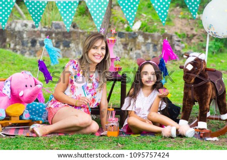 Outdoor view of mom and her daugher sitting in the grass, little girl with a cupcake in the hand, surrounding of party arrangement in the garden on sunny summer day