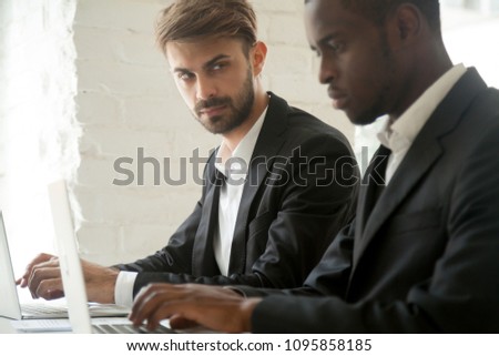 Suspicious cunning male Caucasian worker looking at serious working African American colleague, feeling mad and sneaky distrusting, having doubts, planning. Concept of office relationships, jealousy Royalty-Free Stock Photo #1095858185