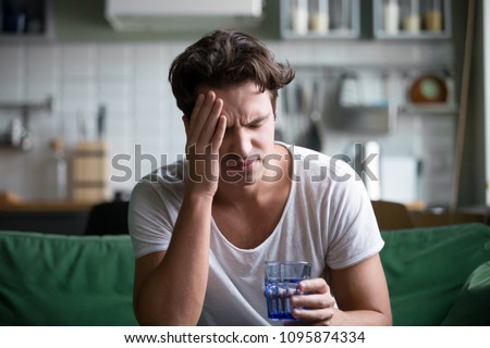 Young man suffering from strong headache or migraine sitting with glass of water in the kitchen, millennial guy feeling intoxication and pain touching aching head, morning after hangover concept Royalty-Free Stock Photo #1095874334