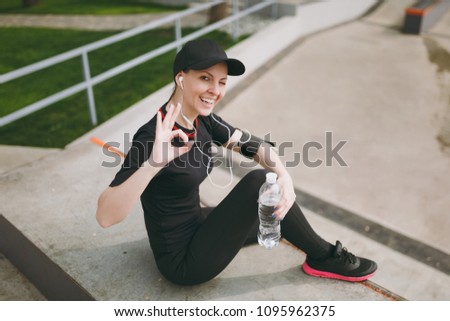 Young athletic smiling girl in black uniform, cap with headphones listening music, holding bottle with water, sitting on training, showing OK sign in city park outdoors. Fitness, healthy lifestyle