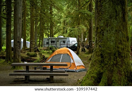 The Campground - Small Orange Tent and Travel Trailer in the Background. Deep Forest Campground. Outdoor Lifestyle Photo Collection.