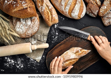 Woman hands cutting a loaf of bread on rustic wooden board, with wheat ears and knife, top view.