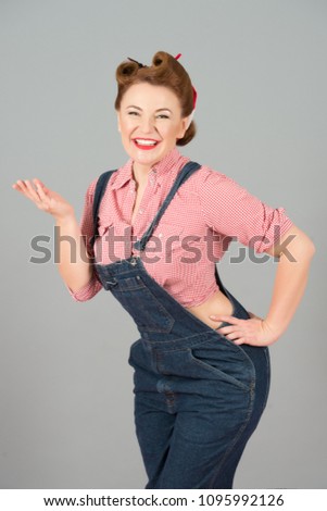 Portrait of smiled and cheerful pin-up girl standing with arm on hips and holding copy space on her palm isolated over gray background. Brunette happy woman in denim with open palm hand.