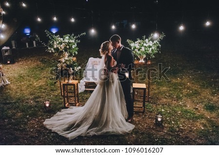 Fashionable newlyweds stand at night against the backdrop of chic decorations and flowers,electric bulbs and garlands,in the backlight. Night shooting of bride and groom.Floristics. Grain,film effect  Royalty-Free Stock Photo #1096016207