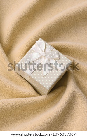 A small gift box in orange with a small bow lies on a blanket of soft and furry light orange fleece fabric with a lot of relief folds. Packing for a gift to your lovely girlfriend