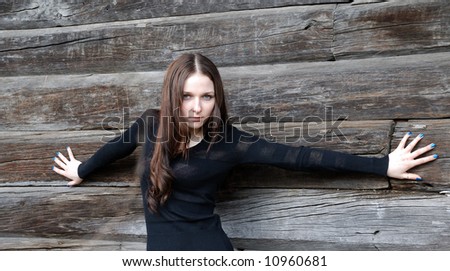 Teenage grave girl in black spreads hands and leans against timber wall.