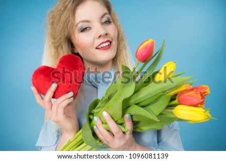 International womens or valentines day. Attractive happy woman blonde hair fashion make up holding tulips bunch and red heart sign. On blue