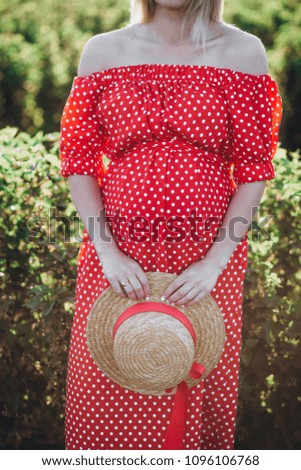 Young beautiful pregnant woman wearing red dress and heat walking with flowers in the green field on a sunny summer day. Nature in the country.