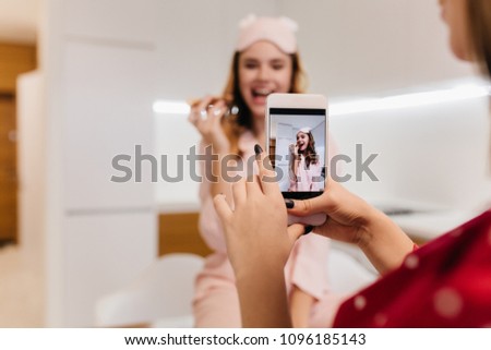 Female model in red attire using phone while taking pictures of friend. Indoor portrait of girl in good mood eating pizza in morning.