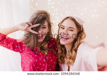 Stunning girl in red night-suit posing with peace sign near sister. Adorable curly lady in eyemask making selfie with her female friend.
