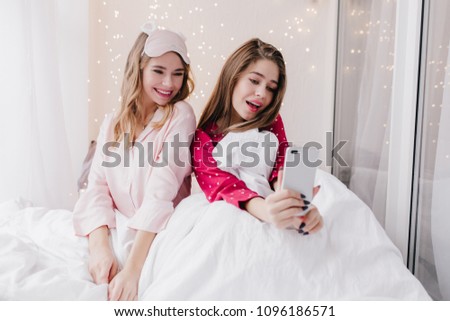 Glamorous young woman in red pajamas making selfie in bed. Dark-haired girl sitting in bedroom with best friend and taking picture of herself.