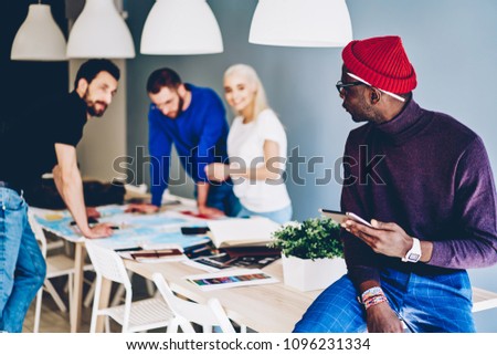 Dark skinned young man communicating with team of colleagues on background while updating software on modern touch pad device using wireless 4G internet.African american hipster guy with tablet