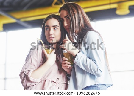 Two beautiful twin girls spend time drinking juice. Sisters relaxing in a cafe and having fun together and whispering.