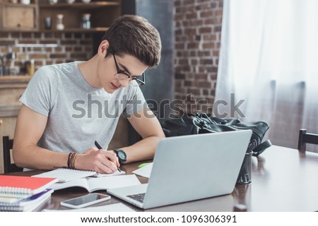 serious student writing homework at table with laptop and smartphone Royalty-Free Stock Photo #1096306391